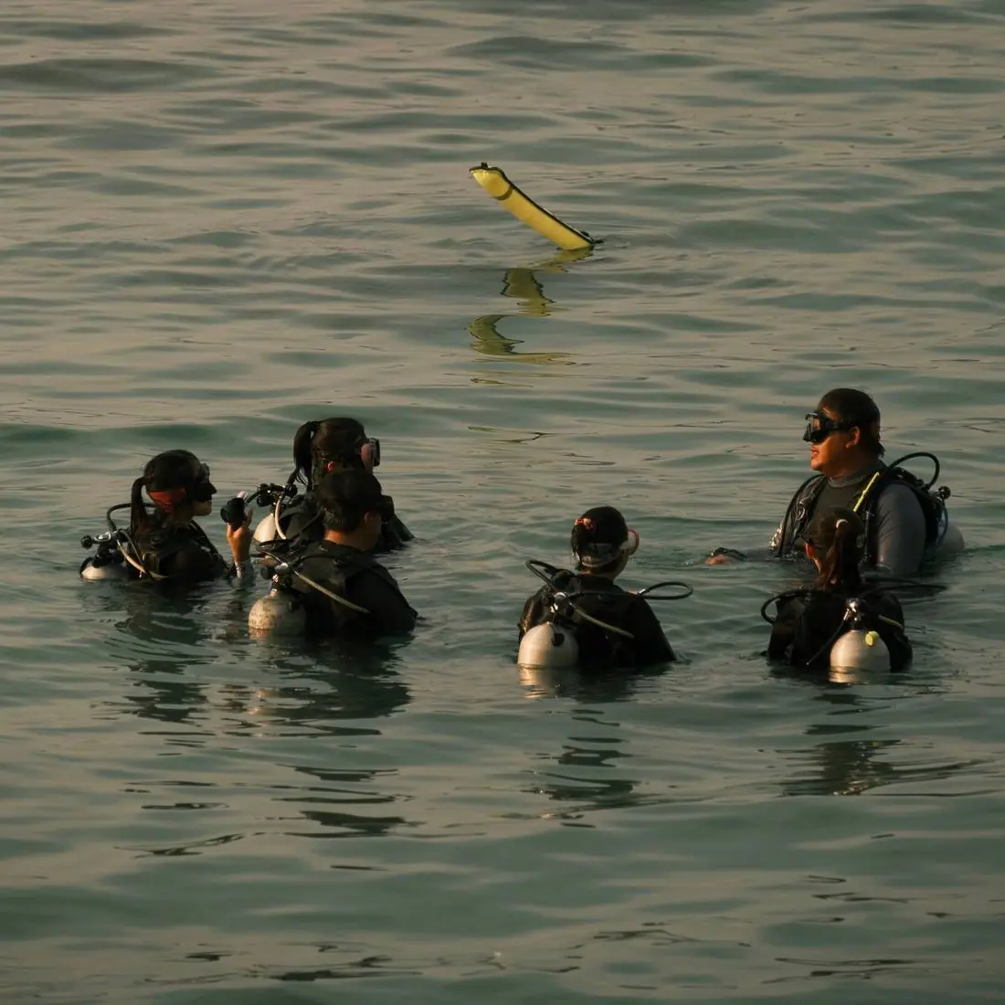 A dive instructor briefing a group of students floating at the surface with a yellow marker buoy